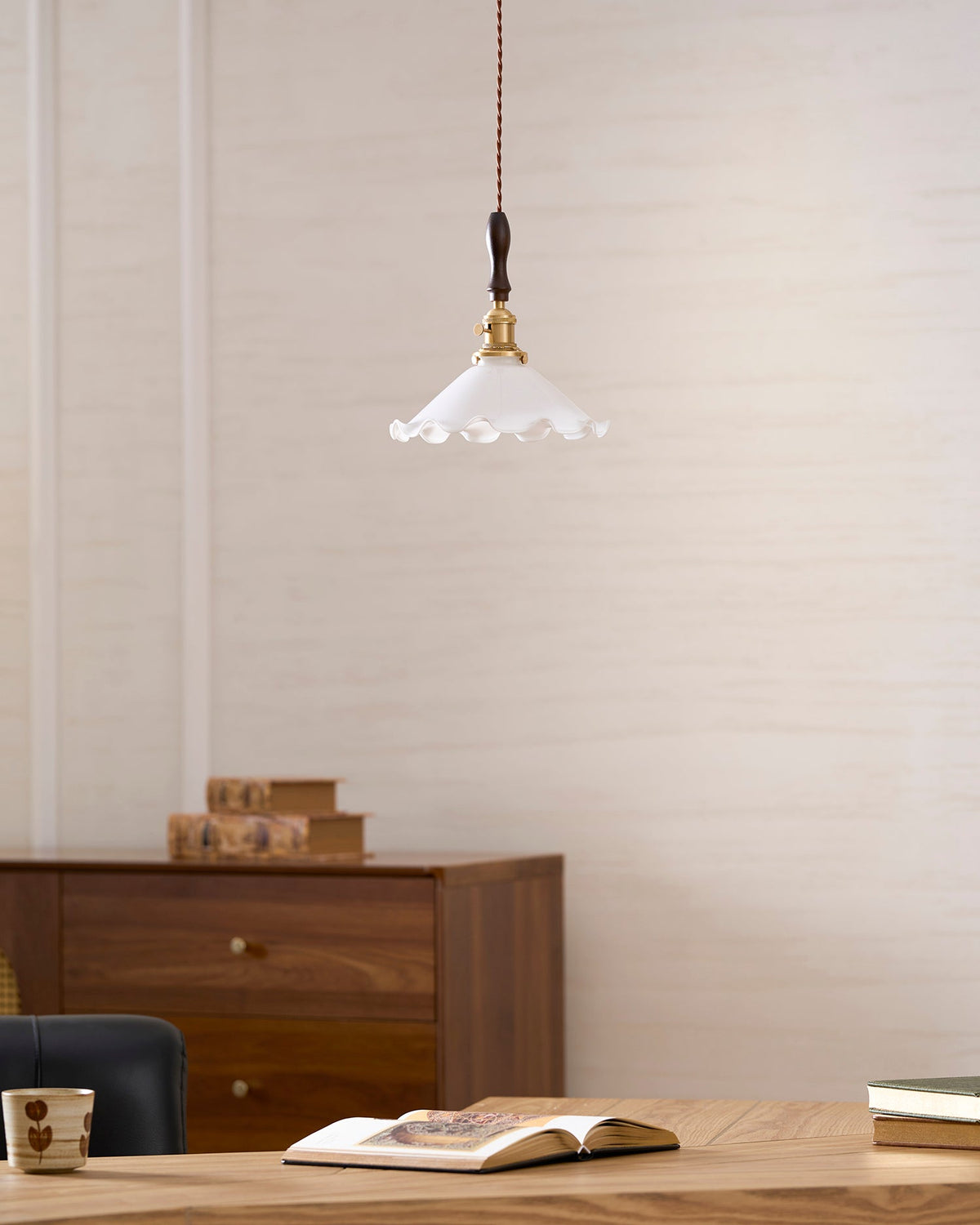 A Clérachic pendant light hangs on a cream wall above a walnut cabinet, an open book and a mug on a wooden desk.