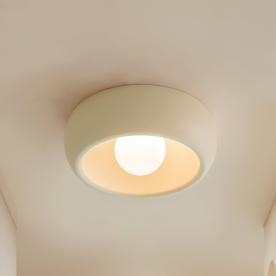 Close view of a beige Clérachic ceiling light mounted on a warm cream ceiling, its round bulb lit inside the rim.