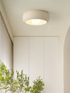 Beige Clérachic ceiling light above a white wardrobe wall and tall plants, seen from the floor of a bright hallway.