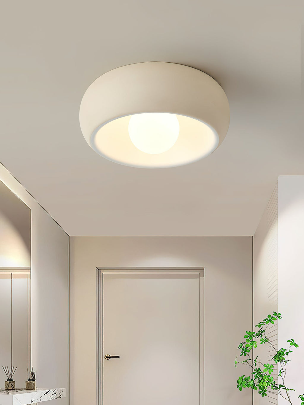 A beige Clérachic ceiling light lit in a hallway with a white door at the far end and a plant at the right.