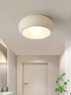 A beige Clérachic ceiling light lit in a hallway with a white door at the far end and a plant at the right.