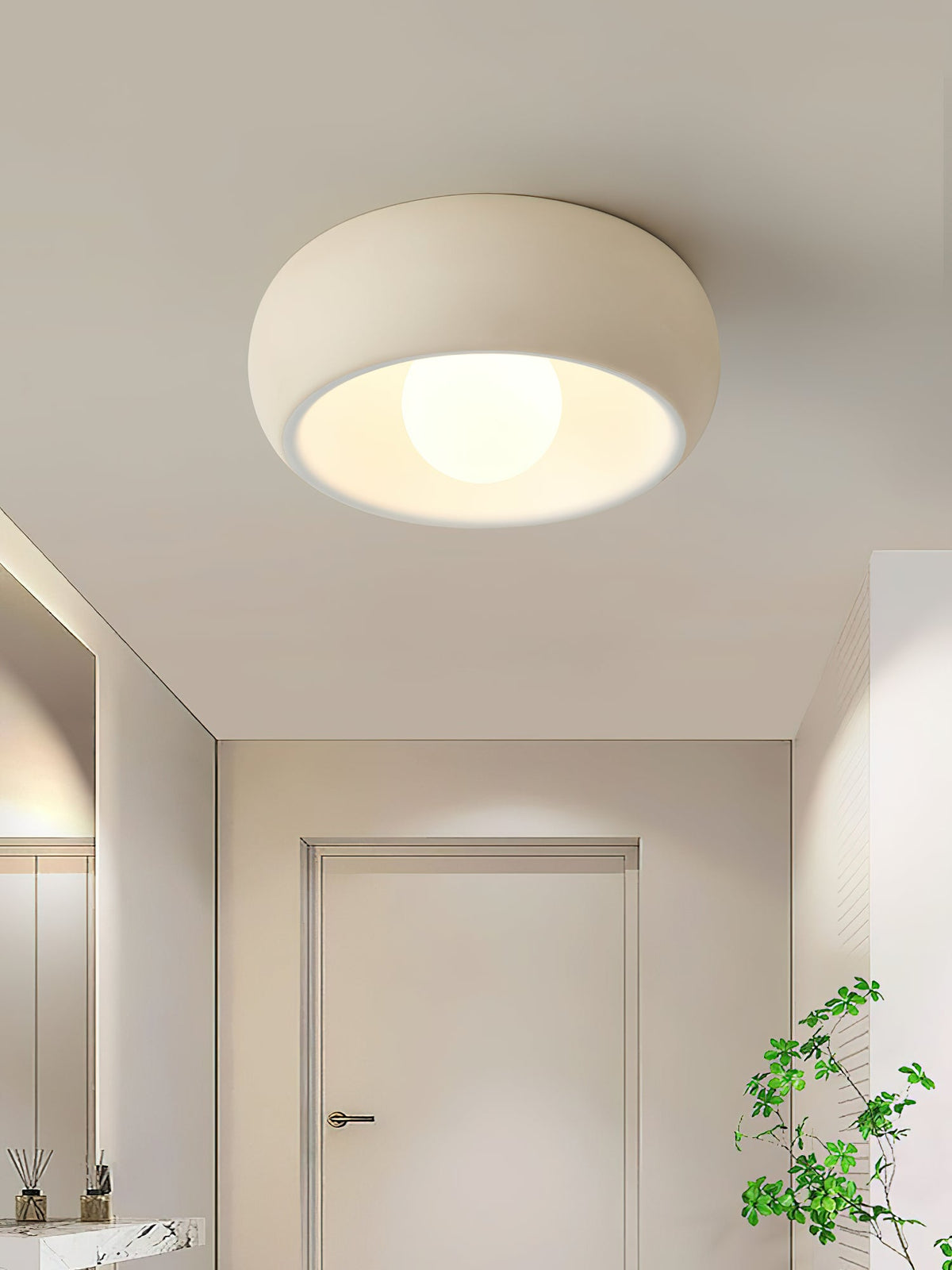 A beige Clérachic ceiling light lit in a hallway with a white door at the far end and a plant at the right.