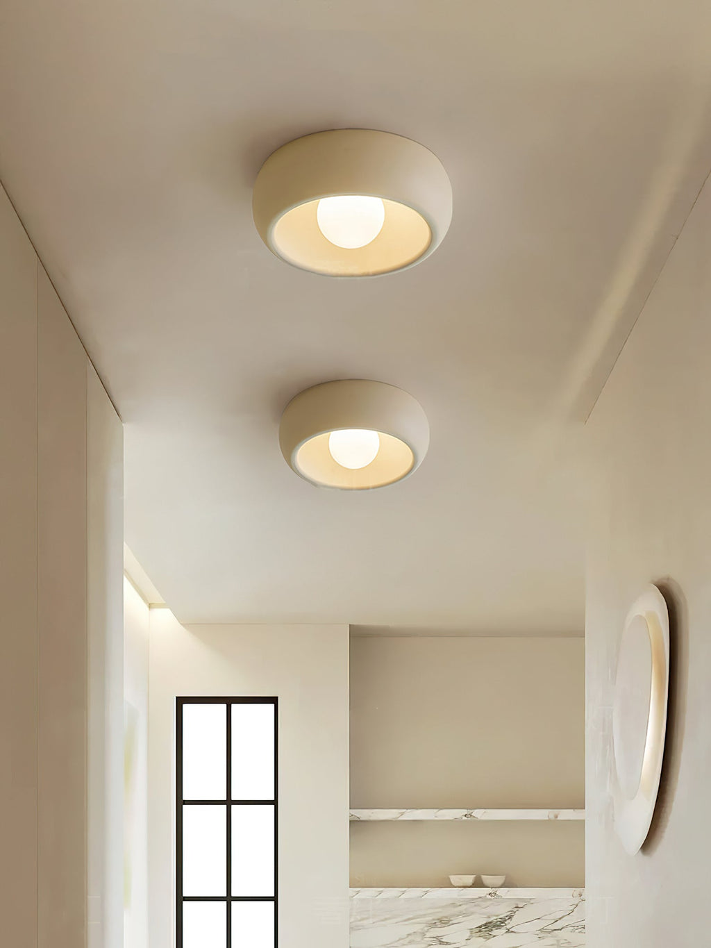 Two beige Clérachic ceiling lights in a row down a pale hallway, which ends in a marble niche and an oval mirror.