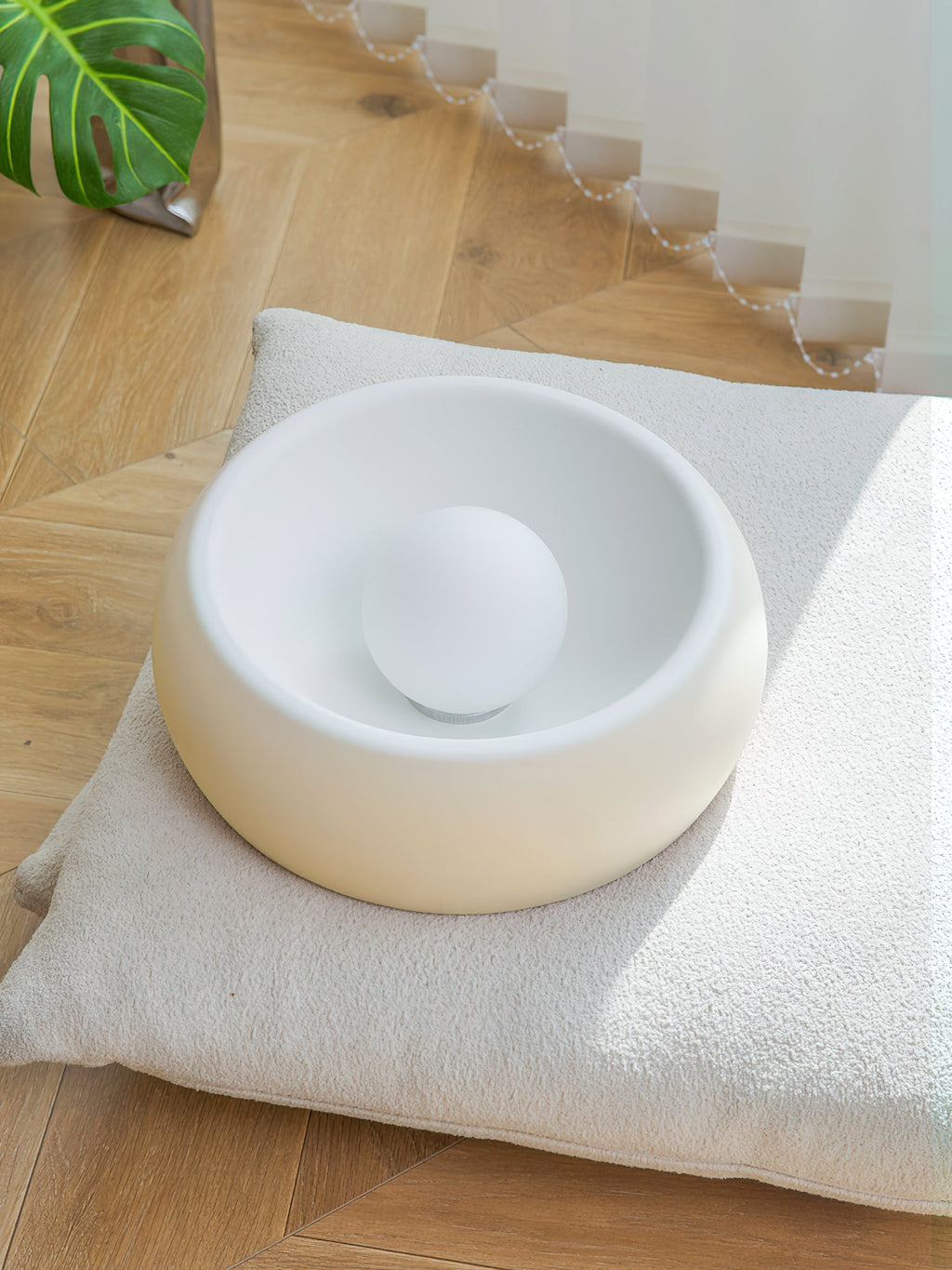 Beige-white Clérachic light rested on a cushion on a wood floor, showing its deep bowl and the round bulb sitting inside.