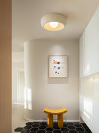 A beige Clérachic ceiling light above a rounded white alcove with a framed print, a yellow stool and black hexagon floor tiles.