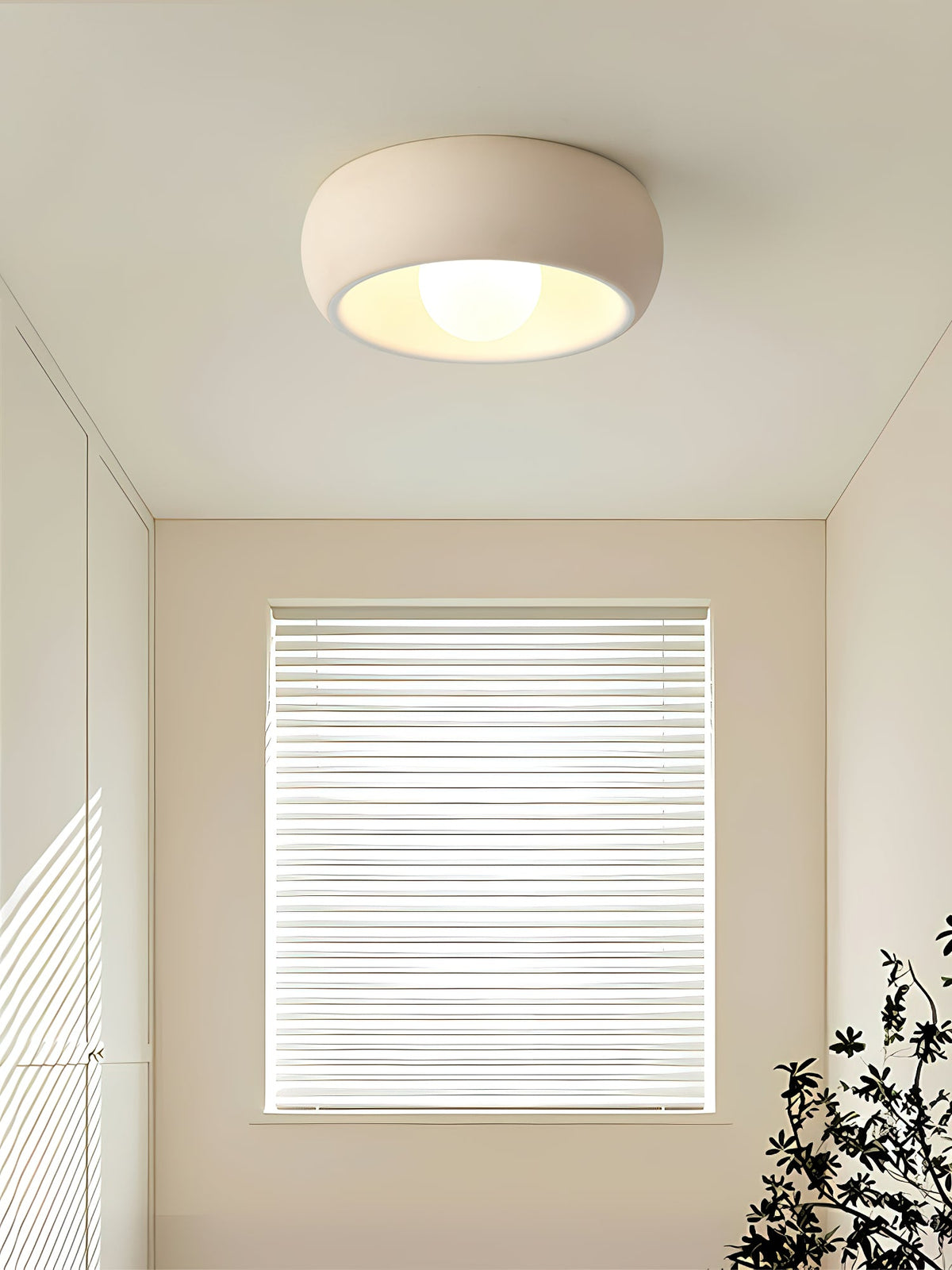 Beige Clérachic ceiling light in a bare, bright room with a window covered by white blinds and a plant at the corner.