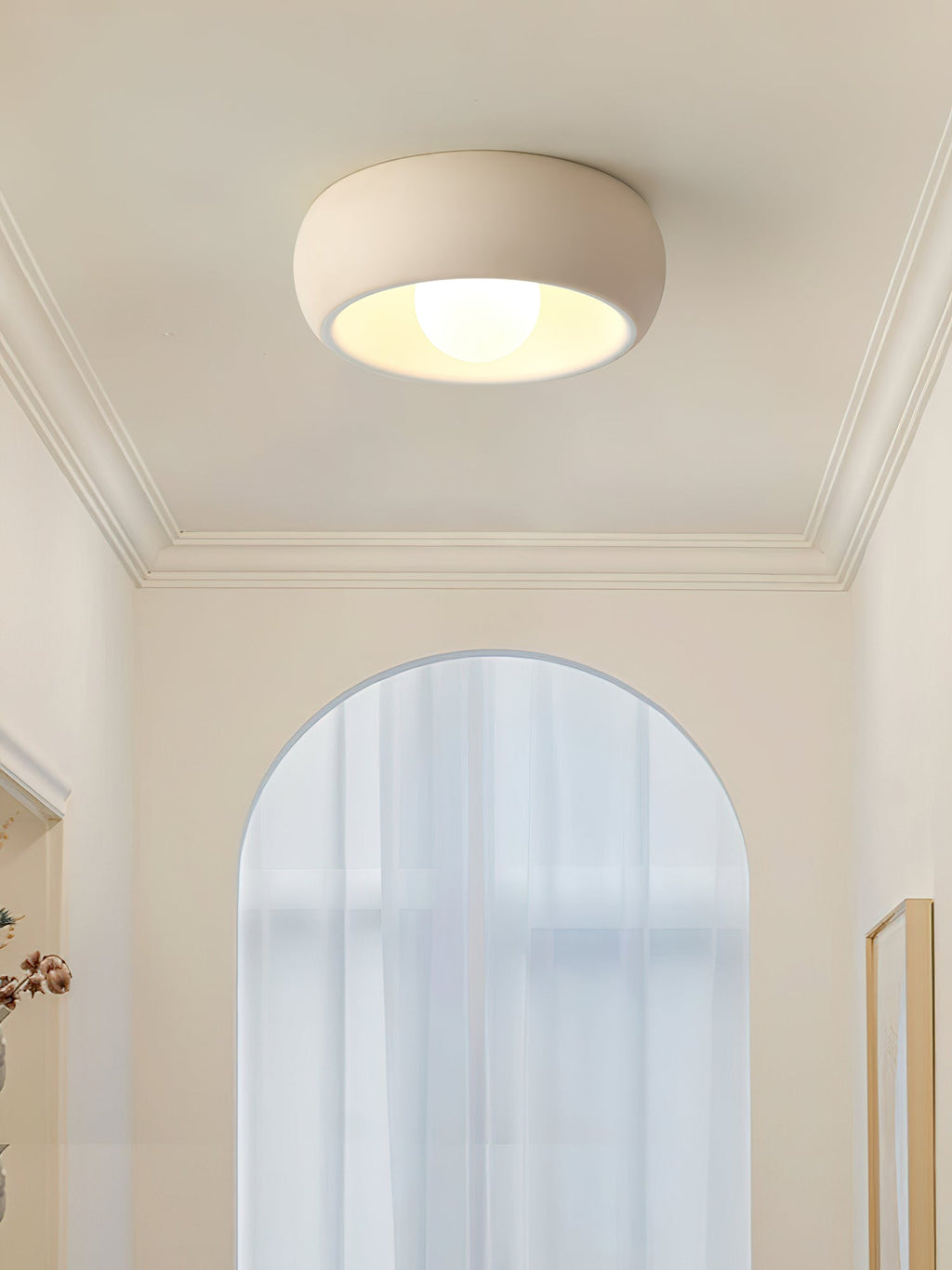 A beige Clérachic ceiling light below a coved ceiling, set against a tall arched window and a pale cream wall.