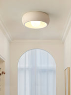 A beige Clérachic ceiling light below a coved ceiling, set against a tall arched window and a pale cream wall.