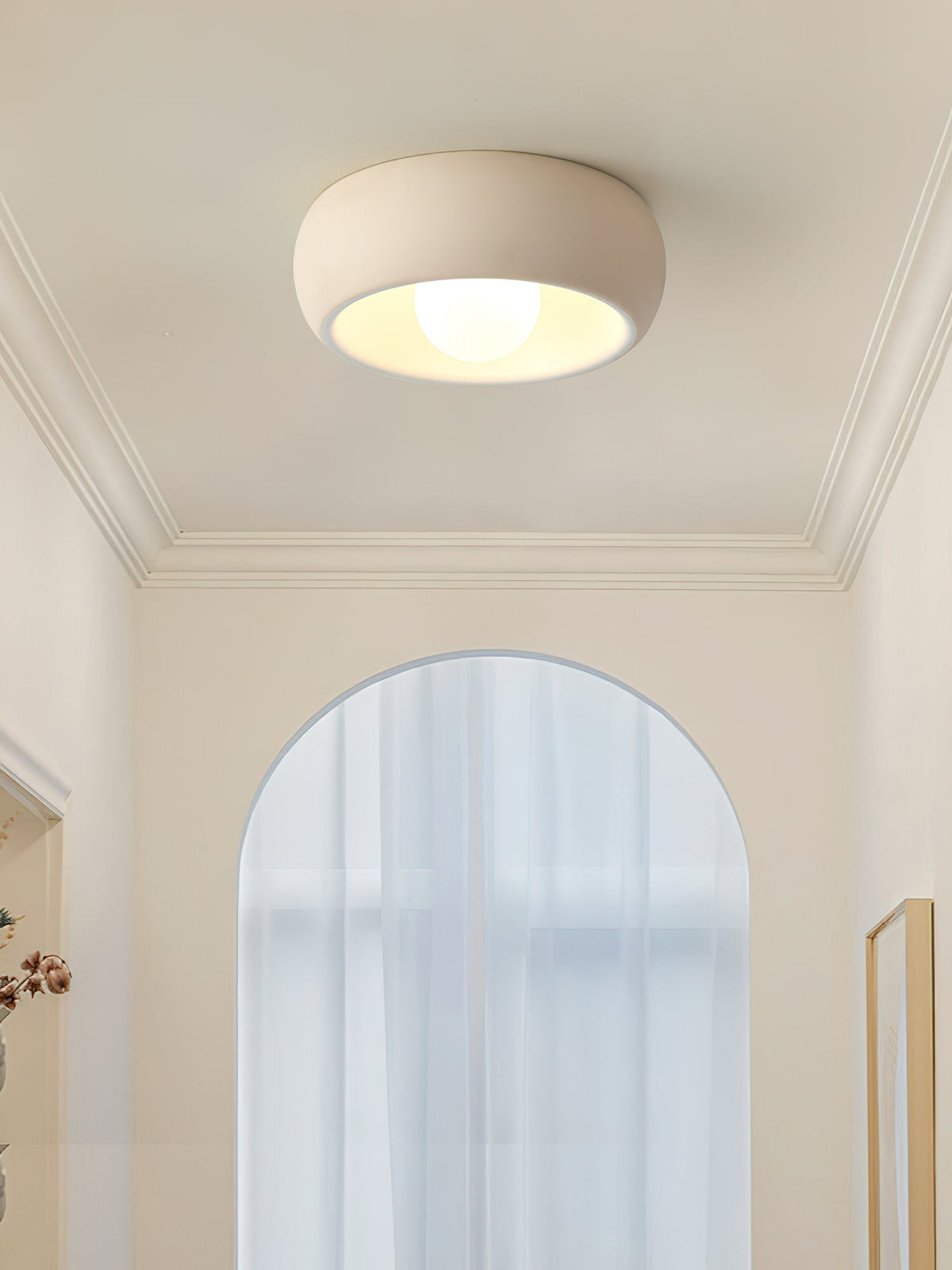 A beige Clérachic ceiling light below a coved ceiling, set against a tall arched window and a pale cream wall.