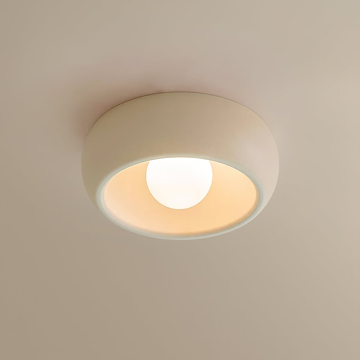Beige Clérachic ceiling light close up on a warm cream ceiling, its pale bowl glowing around the round bulb.