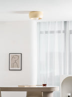 A beige Clérachic ceiling light high in a white room, above a long table, sheer curtains and a framed line drawing.