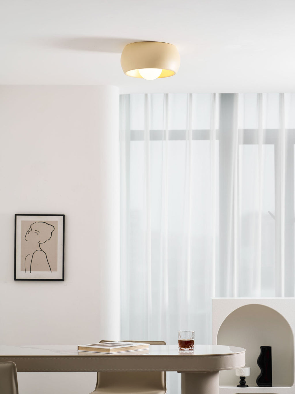 Beige Clérachic ceiling light in a white dining room, glowing above an oval table with a glass and a book on top.