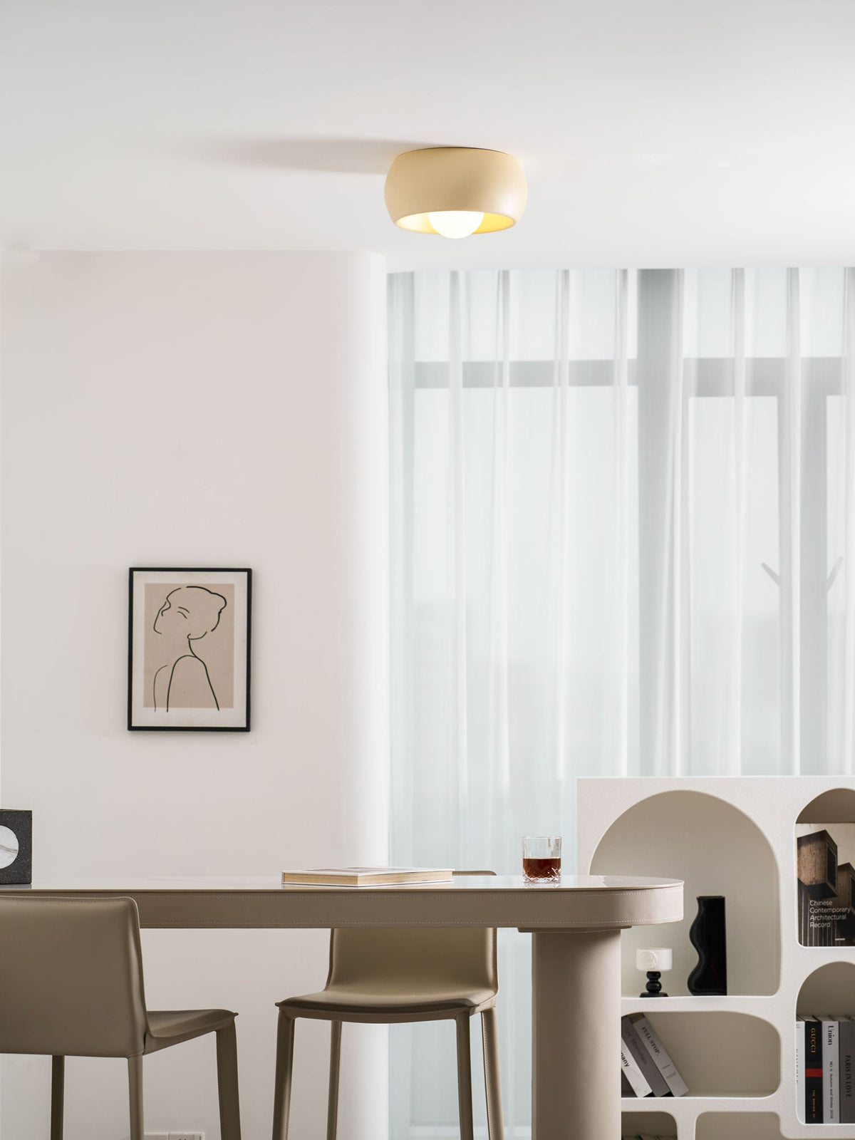 Beige Clérachic ceiling light above a dining table with leather chairs, a framed line drawing and an arched white shelf.