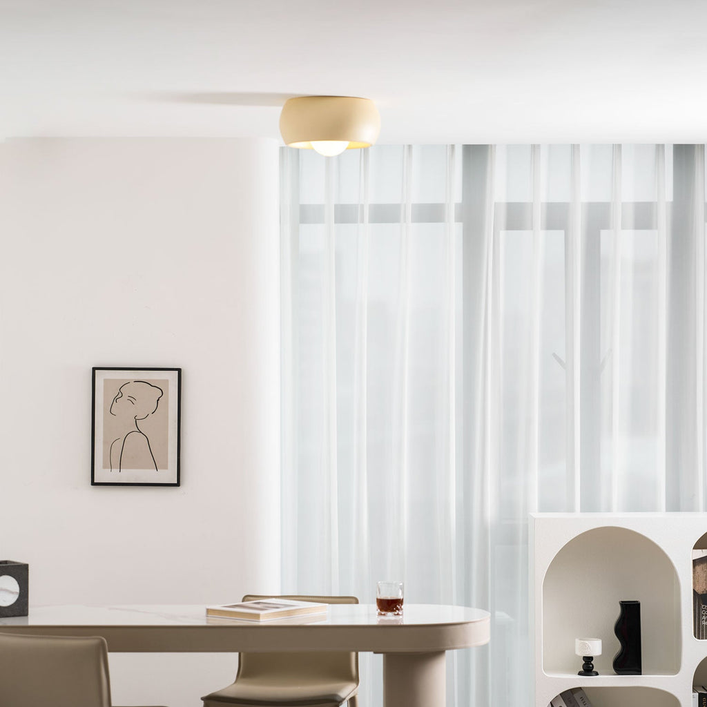 A beige Clérachic ceiling light seen from a distance, over a dining table with grey-green chairs and a tall window of sheers.