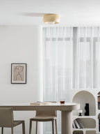 Beige Clérachic ceiling light at the top of a bright dining room, seen above an oval table, a drawing and an arched shelf.