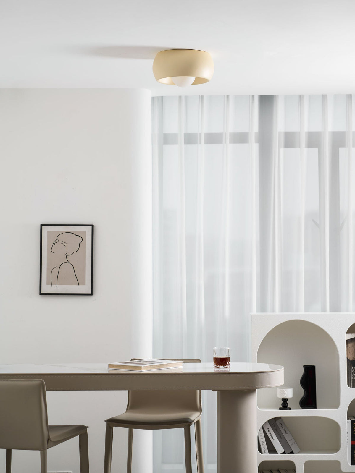 Beige Clérachic ceiling light at the top of a bright dining room, seen above an oval table, a drawing and an arched shelf.