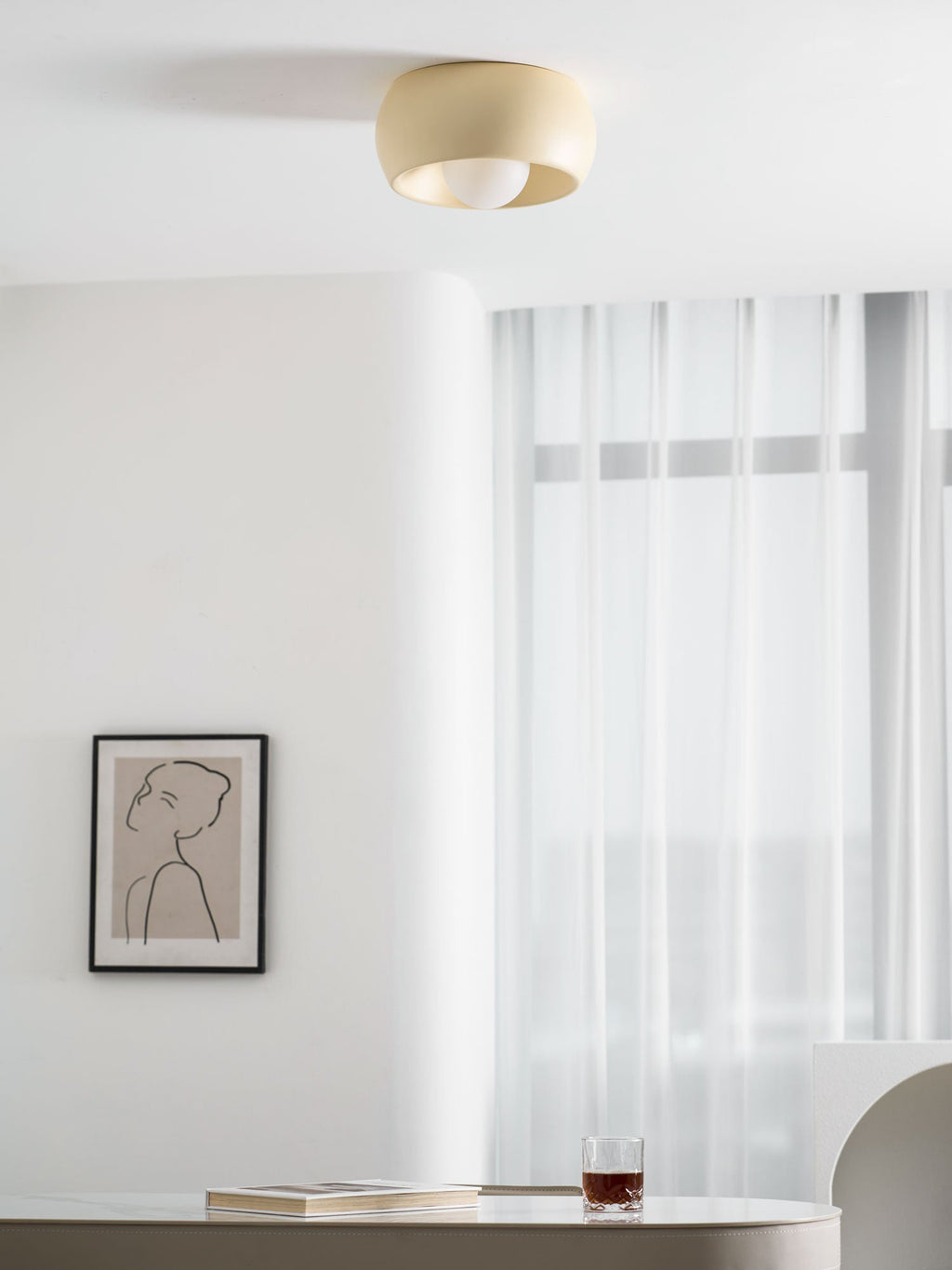 Beige Clérachic ceiling light with its bulb showing, hung above a pale wall with a framed drawing and sheer curtains.