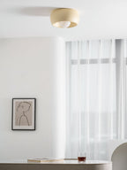 Beige Clérachic ceiling light with its bulb showing, hung above a pale wall with a framed drawing and sheer curtains.