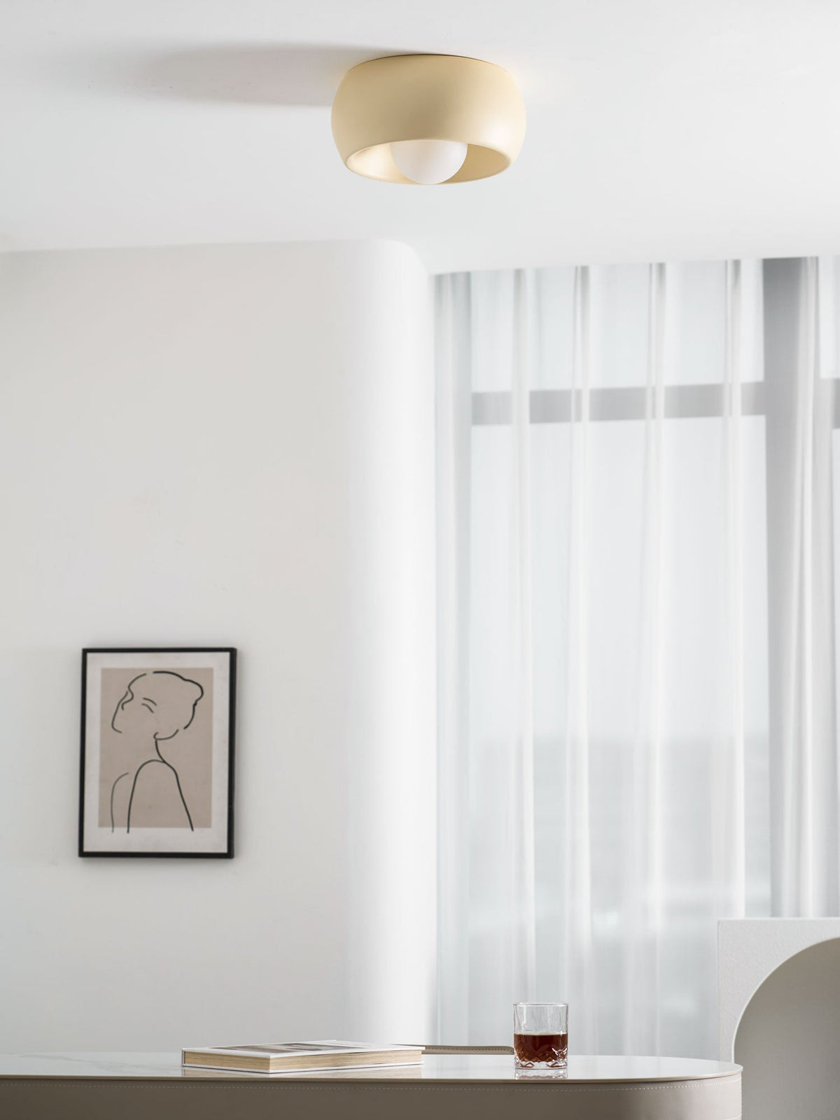 Beige Clérachic ceiling light with its bulb showing, hung above a pale wall with a framed drawing and sheer curtains.