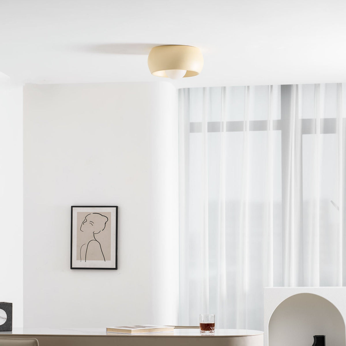 Clérachic ceiling light in beige above a drinks glass on a table, with a framed line drawing and sheer curtains behind.