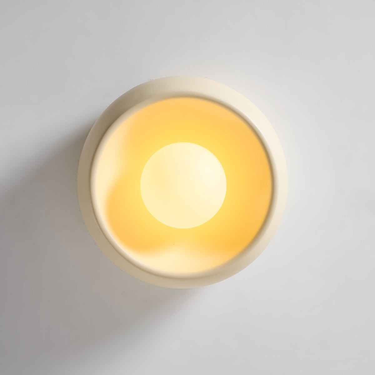 Face-on view of this beige Clérachic ceiling light on a pale grey surface, with a warm glow inside its bowl around the round bulb.