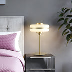 A lit Clérachic table lamp with a white glass shade and gold stem on a dark bedside table, beside a pink quilted bed and a plant.