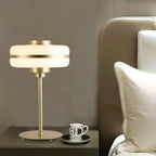 This Clérachic table lamp, with a lit white glass shade and a gold base, stands beside a teacup on a bedside table and a cushioned headboard.