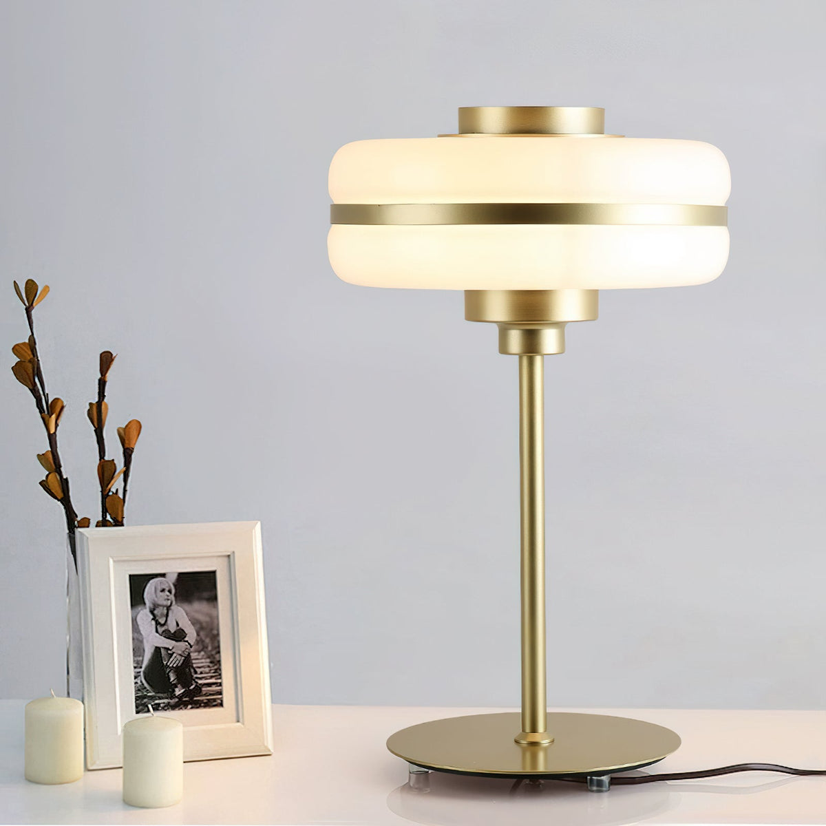 Clérachic table lamp on a white console beside a framed photo, small candles and bare branches, its white shade and gold stem in view.
