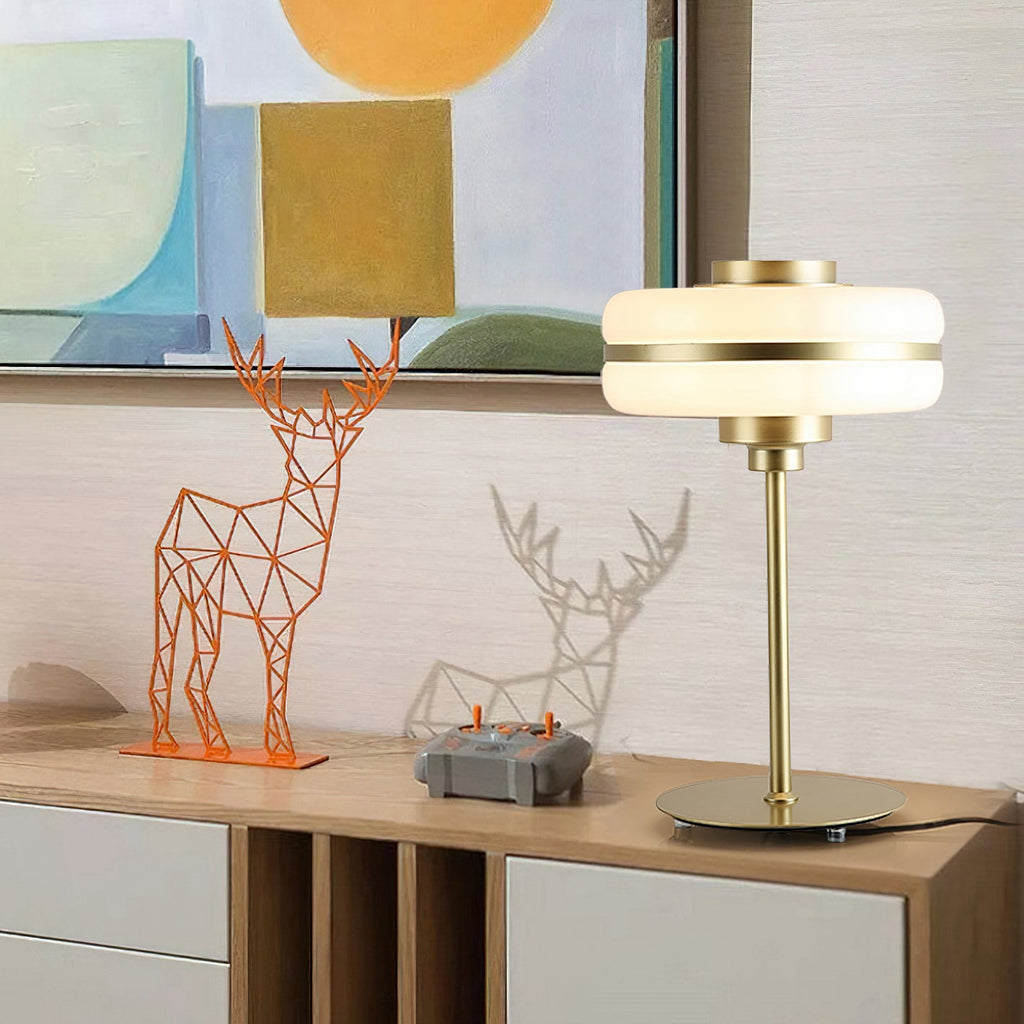 A Clérachic table lamp with a lit white glass shade on a wooden sideboard, beside geometric deer figures and a colourful painting.