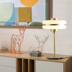 A Clérachic table lamp with a lit white glass shade on a wooden sideboard, beside geometric deer figures and a colourful painting.