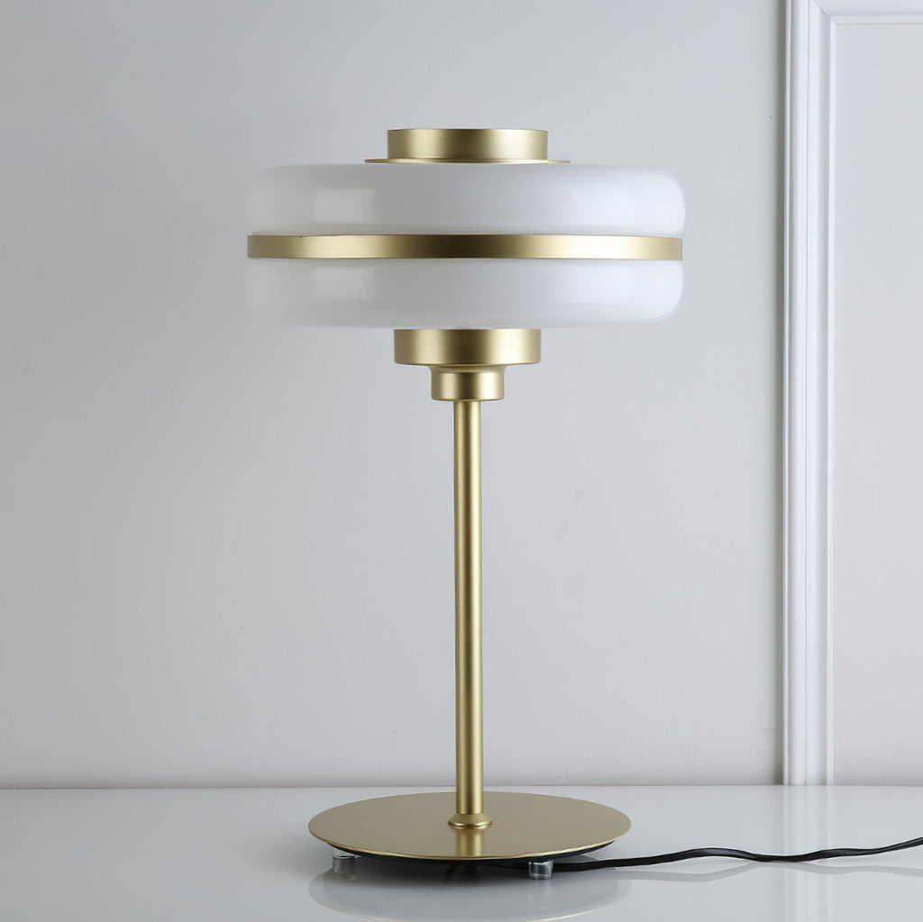 Unlit white glass shade with a gold band on a slim gold stem, this Clérachic table lamp on a white surface against a pale panelled wall.