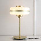 Lit white glass shade with a gold band on this Clérachic table lamp, set on a white surface with its black cord trailing out of frame.