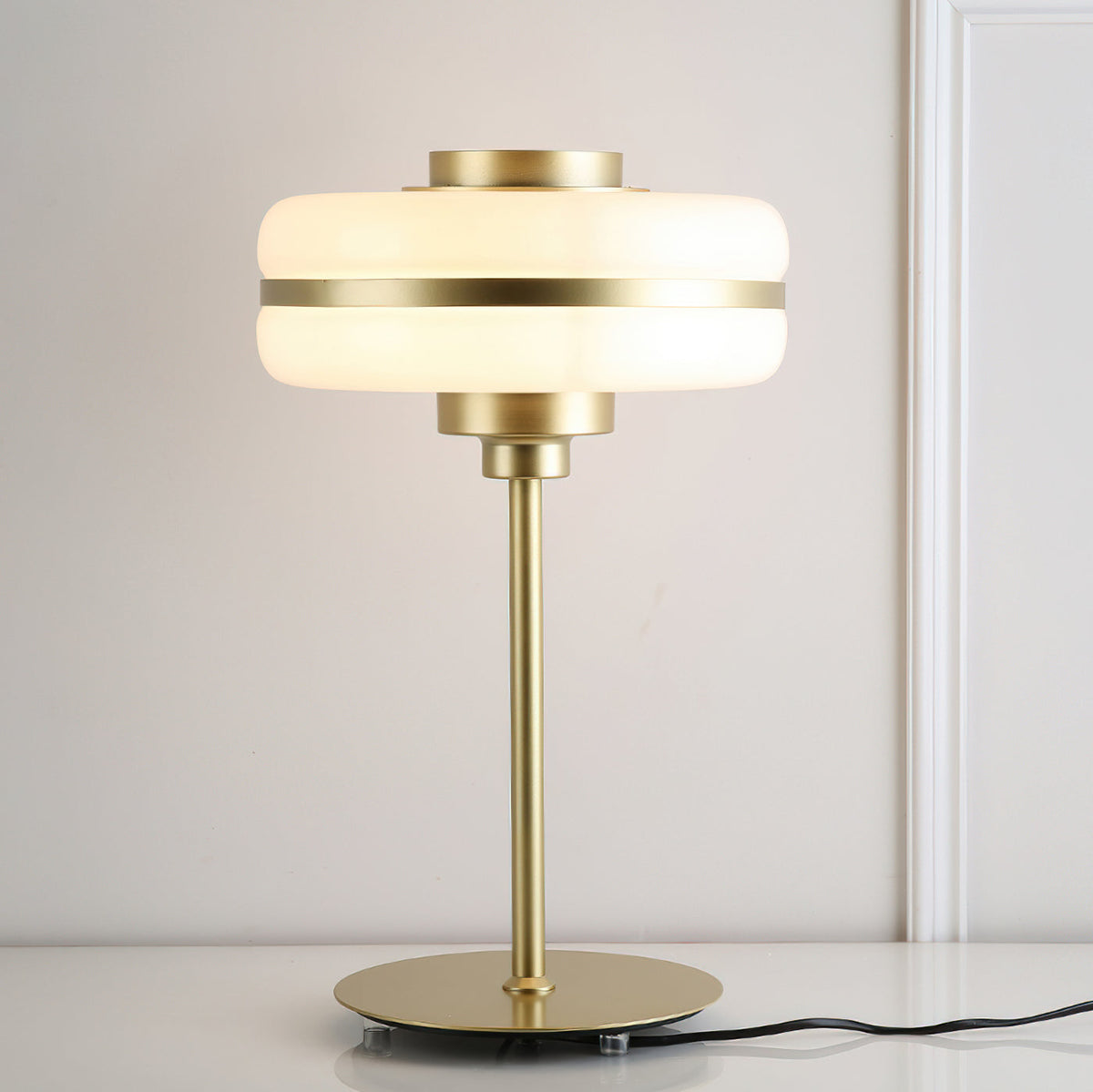 Lit white glass shade with a gold band on this Clérachic table lamp, set on a white surface with its black cord trailing out of frame.
