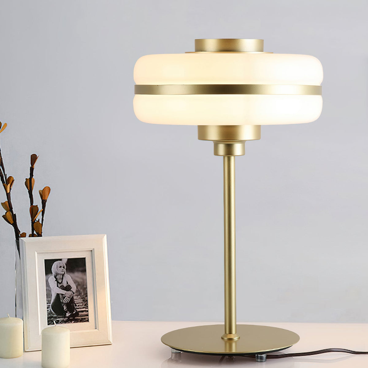 Clérachic table lamp lit beside a framed black and white photo and candles, its gold stem and round base clear against a grey wall.