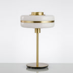 Unlit Clérachic table lamp with a white glass shade, gold band and slim gold stem on a pale surface against a soft grey backdrop.