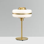 This Clérachic table lamp with an unlit white glass shade and a gold stem and round base stands against a plain grey wall.