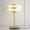 Lit white glass shade with a gold band on a slim gold stem, this Clérachic table lamp in front of a white panelled wall.