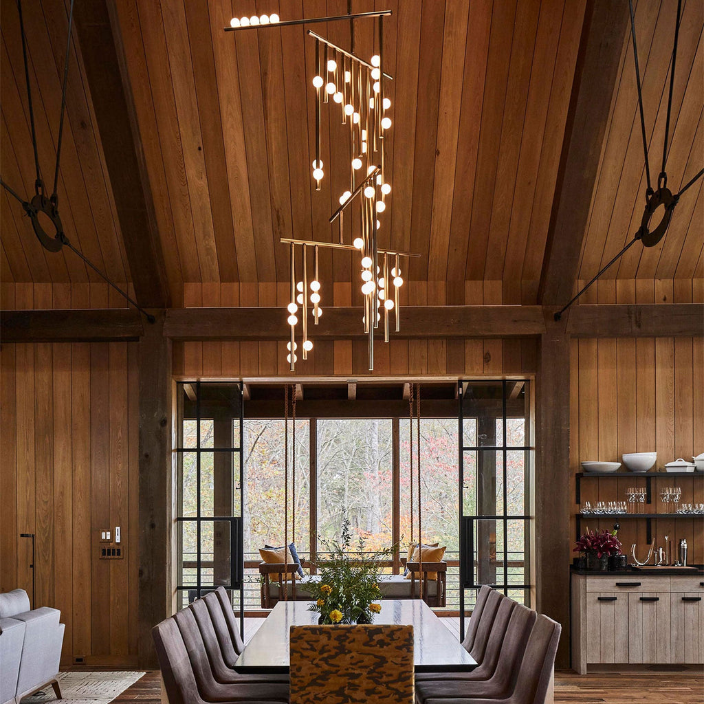 A Clérachic chandelier hanging in a timber-lined dining hall under a pitched ceiling, its lit orbs spread over several brass bars above a long table.