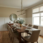 Above a walnut dining table in a beige room, a Clérachic pendant of brass bars carries white glass orbs at staggered heights.