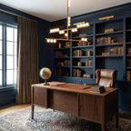 Brass horizontal bars with white orbs on a Clérachic pendant, hung above a walnut desk in a study with dark blue bookshelves.