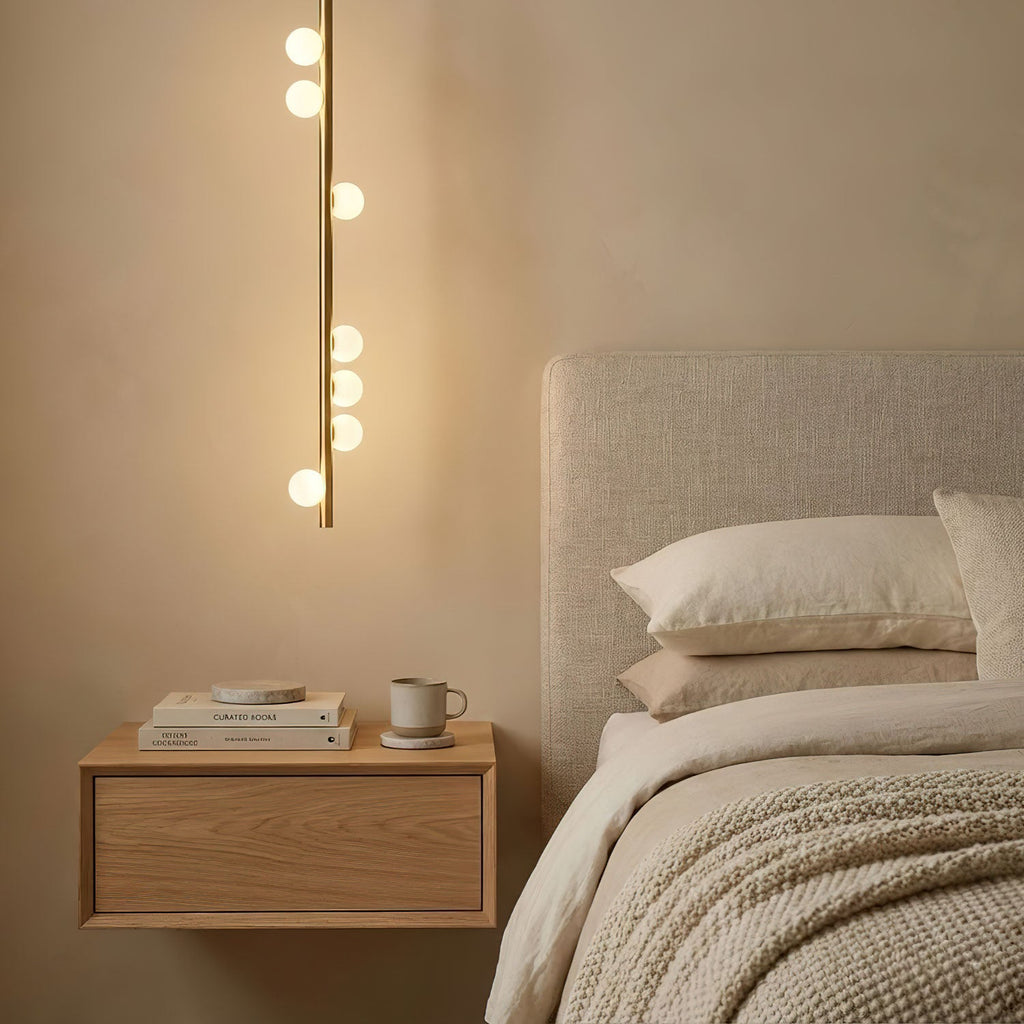 A single vertical brass rod of this Clérachic pendant, with white glass orbs along it, hangs beside a bedside shelf and a cream headboard.