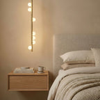 A single vertical brass rod of this Clérachic pendant, with white glass orbs along it, hangs beside a bedside shelf and a cream headboard.