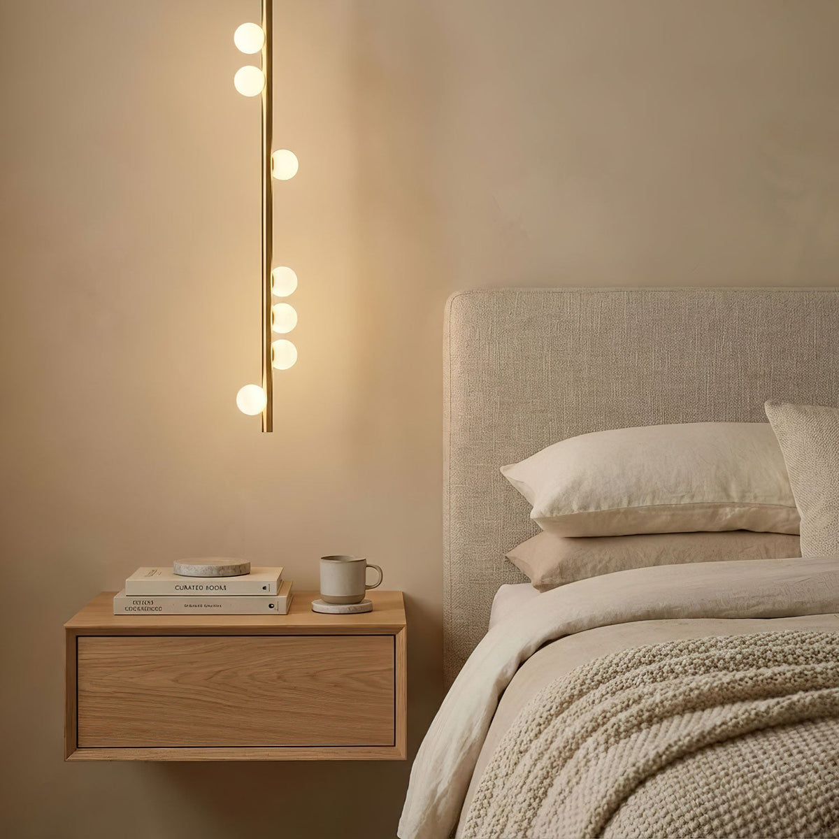 A single vertical brass rod of this Clérachic pendant, with white glass orbs along it, hangs beside a bedside shelf and a cream headboard.