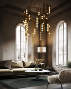 A Clérachic chandelier fills a tall room with arched windows, its brass rods and lit orbs hanging low over a green rug and a low sofa.