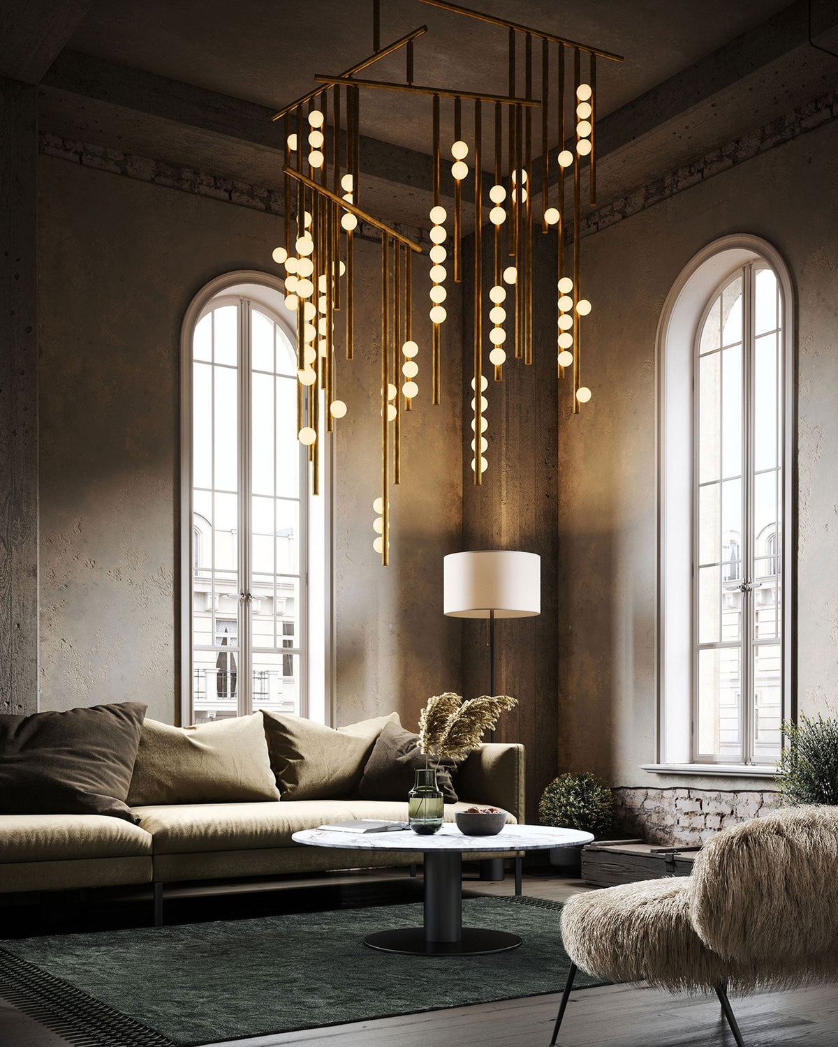 A Clérachic chandelier fills a tall room with arched windows, its brass rods and lit orbs hanging low over a green rug and a low sofa.