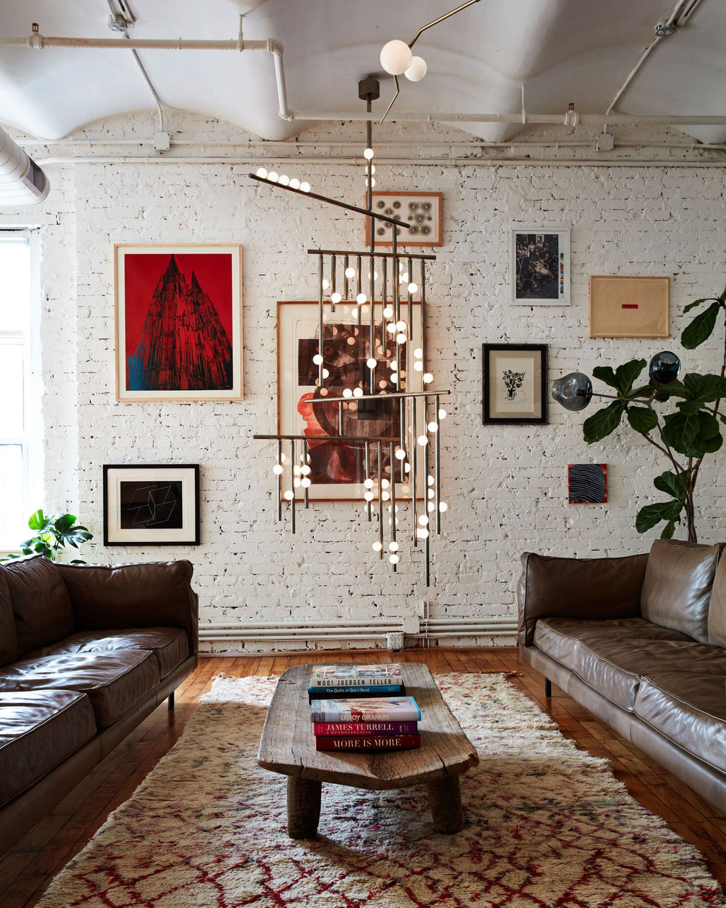 Brass rods and white orbs of a Clérachic chandelier hang in front of a white brick wall hung with art, above a leather sofa.