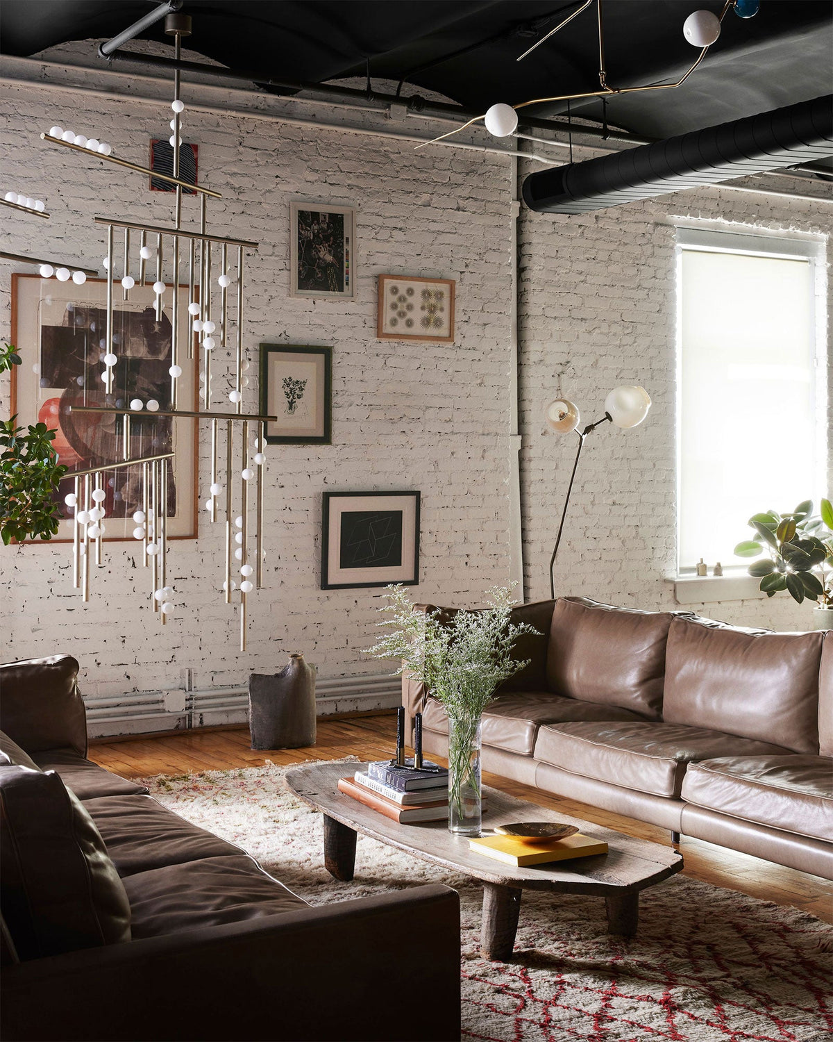 In a loft with exposed ducts and brick walls, a Clérachic chandelier of fine rods and white orbs hangs above a low wooden coffee table.