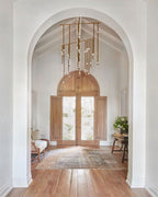 A Clérachic chandelier hangs in an arched white entrance hall with double timber doors, its brass rods dropping towards a wooden floor.