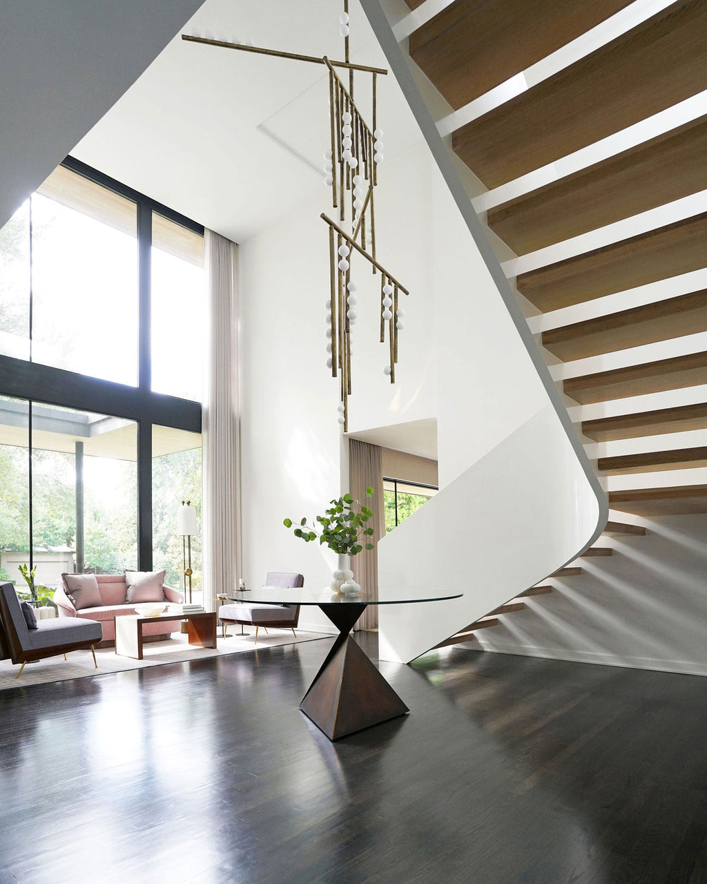 In a double-height room with a floating stair and a glass wall, a Clérachic chandelier hangs over a round table with a dark base.