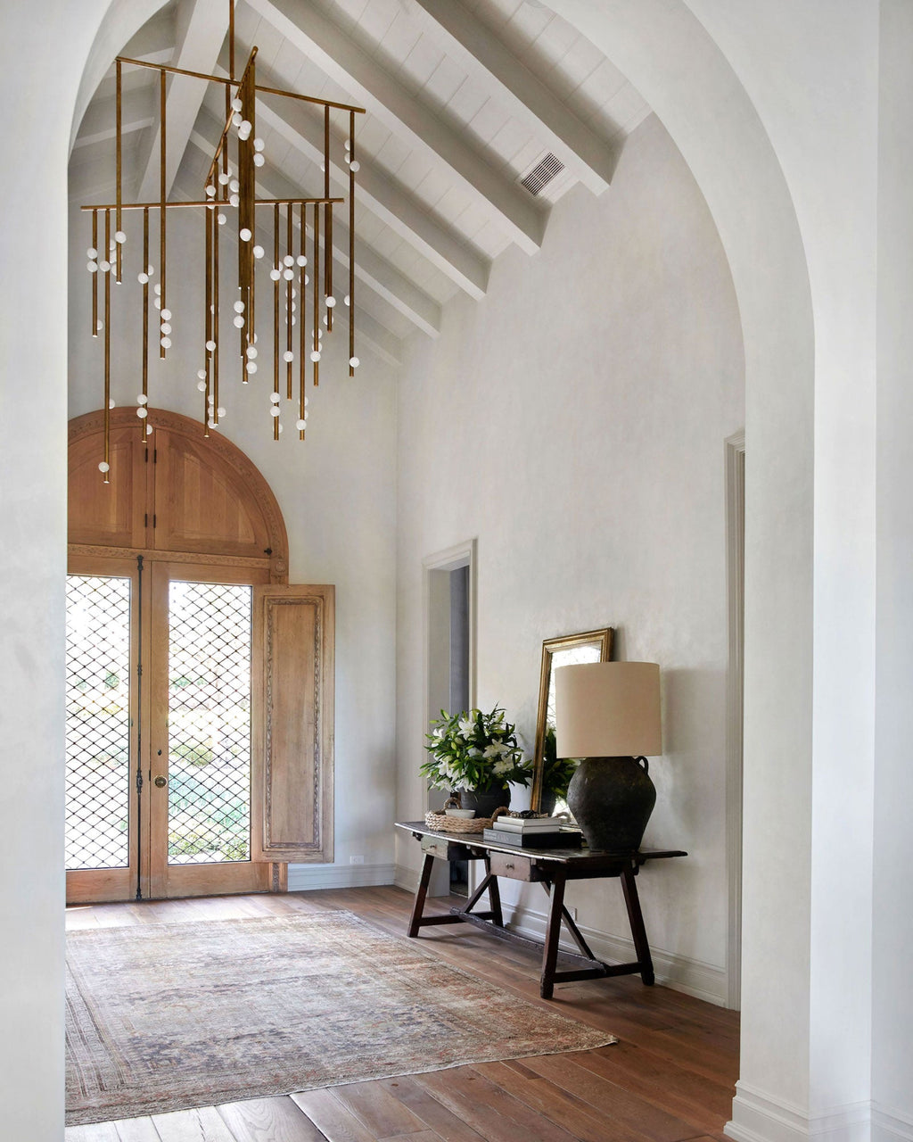 A Clérachic chandelier hangs from a pitched ceiling in an arched entrance hall, with a console table and a lamp by the timber doors.